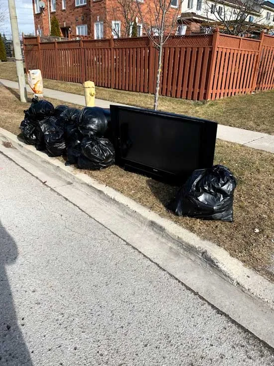 Garbage bags and an old flat-screen TV left at the curb beside a fence